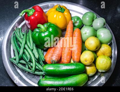 Assorted vegetables of green beans, red bell pepper, yellow bell pepper, green bell pepper, carrots, cucumbers, green tomatoes, and lemons on a plate Stock Photo