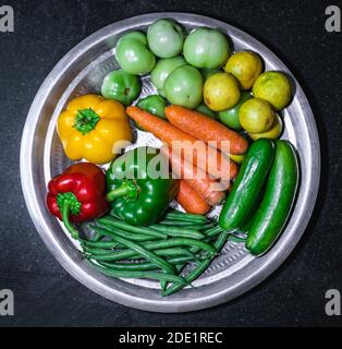Green beans, red bell pepper, yellow bell pepper, green bell pepper, carrots, cucumbers, green tomatoes, and lemons on a stainless steel plate Stock Photo