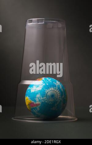A toy in the shape of a globe is covered with a plastic glass. Concept on the topic of clogging the planet with plastic. Stock Photo