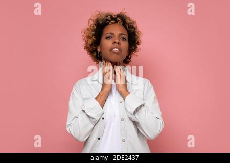 Young african american woman with curly hair touching painful neck, sore throat for flu. Studio shot on pink wall. Stock Photo
