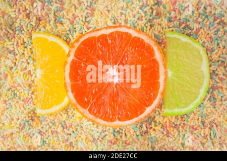Fun and colorful composition of half slices of orange and lime and a large grapefruit on a background of colored sweets. Fruit and sugar whims. Stock Photo