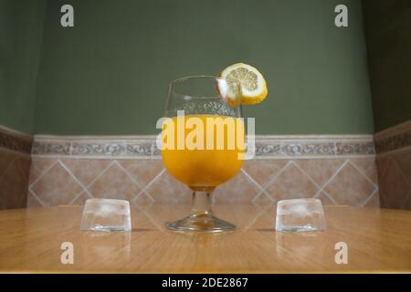 Refreshing orange and apple juice garnished with a lemon slice in a crystal glass and two ice cubes on both sides on a table in a rustic kitchen. Stock Photo