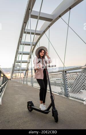 A young beautiful woman is standing next to her electric scooter with one foot on a standing platform. She is wearing a hat and she is business-traine Stock Photo