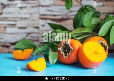 Ripe persimmon with slices on blue table against green leaves Stock Photo