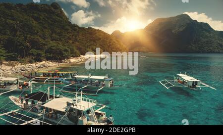 Sun over mountain ranges with green tropic forest at ocean bay. People rest on sand beach near traveling boats. Tourist summer vacation at sunny day on Palawan Island, Philippines, Asia. Aerial view Stock Photo
