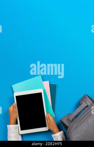 Kid hands with tablet computer, notebooks, books and gray backpack over light sky blue background Stock Photo