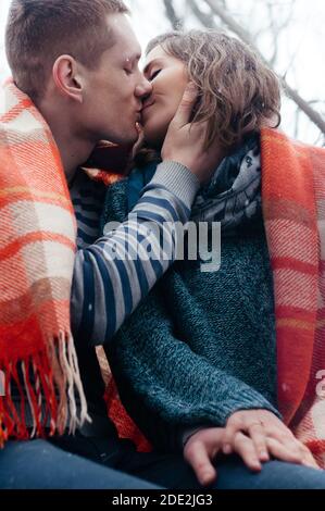 Christmas, season and people concept - happy couple sitting covered with a blanket and kissing holding each other's hand outdoors in winter park Stock Photo