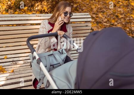 Stylish mom with stroller talking on the phone while sitting on  bench in the park while the child is sleeping Stock Photo