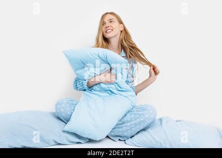 Beautiful girl in pajamas, sitting on the bed with pillows in her hands, on a white background, a woman wakes up in bed Stock Photo