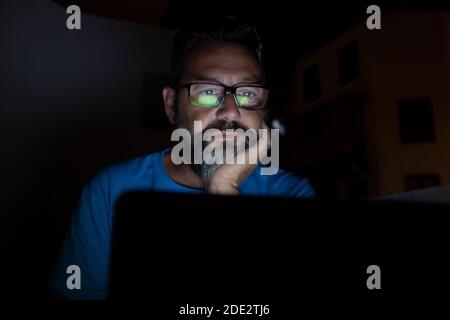 Attractive mature man at home at night while using laptop, light on the face. Bearded man with eyeglasses Stock Photo