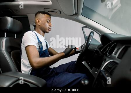 Young African male car service worker in uniform performs a professional vehicle interior cleaning, wiping car steering wheel with a brush at the car Stock Photo