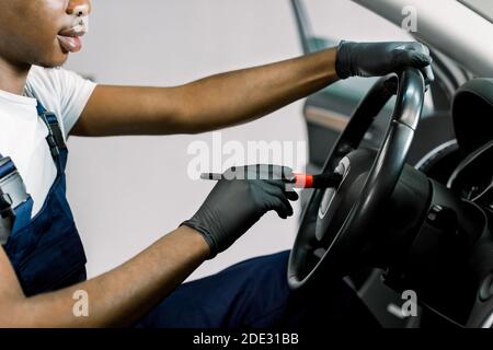 Close-up cropped image of African dark skinned worker performing professional car interior cleaning, wiping steering wheel with a brush, remowing the Stock Photo