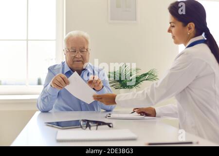 Smiling woman doctor giving paper prescription to elderly man patient in clinic