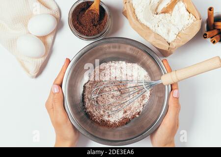 Female hands holding bowl with whisk and powder for baking homemade chocolate cake with cinnamon on white table, top view. Stock Photo
