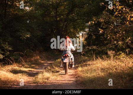 Back view of woman riding motorcycle on dirt road in forest Stock Photo