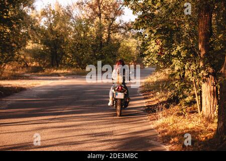 Back view of woman riding motorcycle on country road amidst trees Stock Photo