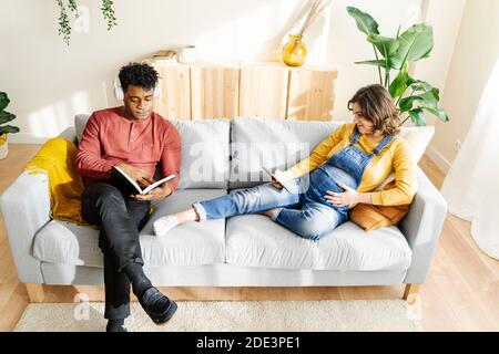 Husband listening to music and reading a book with his pregnant wife who is watching a tablet on a sofa. Interracial couple concept Stock Photo