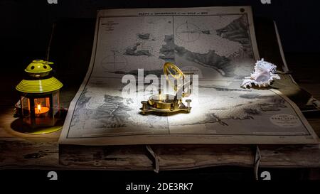 Antique nautical maps, placed on a wooden table, illuminated by a yellow lantern and held in place by a beautiful shell and a brass compass. Stock Photo