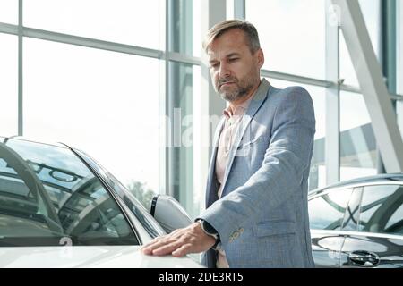 Contemporary mature man in smart casualwear keeping hand on front of new car of white color and looking at it while choosing vehicle Stock Photo