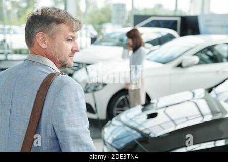 Back view of serious contemporary mature man standing in front of luxurious black car in large auto center with his wife on background Stock Photo