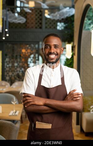 Young waiter of African ethnicity in brown uniform crossing arms on chest while standing in aisle between tables served for guests of cafe Stock Photo