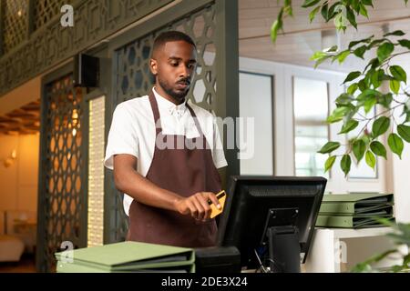 Young waiter or cashier in shirt and apron standing in front of computer and registering payment of client for the ordered food and drink Stock Photo