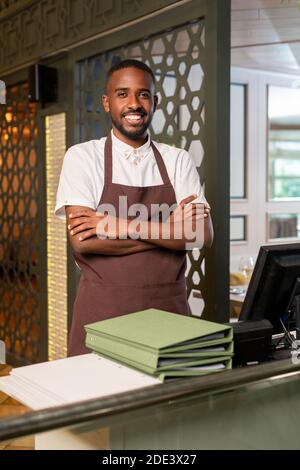 Young cheerful waiter of African ethnicity looking at you with toothy smile and crossing arms on chest while standing in restaurant environment Stock Photo