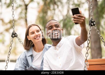Young cheerful intercultural couple making selfie while sitting on bench in park in the morning and looking in smartphone camera with smiles Stock Photo