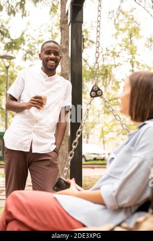 Happy African guy with drink standing in front of young woman relaxing on swing while talking to her during chill on summer day in park Stock Photo