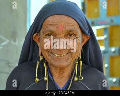 Old Indian Gujarati Rabari woman with face wrinkles and tribal snake earrings (nagali) in her elongated ear lobes smiles at the camera. Stock Photo