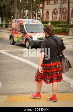 Rear view of young thick man with beard and messenger bag, wearing red plaid skirt with red converse high top tennis shoes approaching crosswalk in do Stock Photo