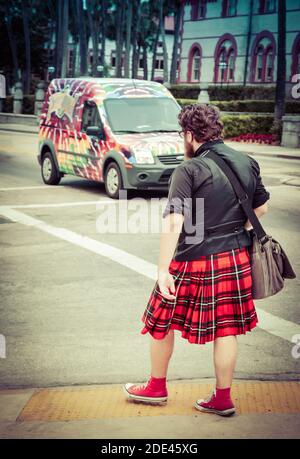 Rear view of young thick man with beard and messenger bag, wearing red plaid skirt with red converse high top tennis shoes approaching crosswalk in do Stock Photo