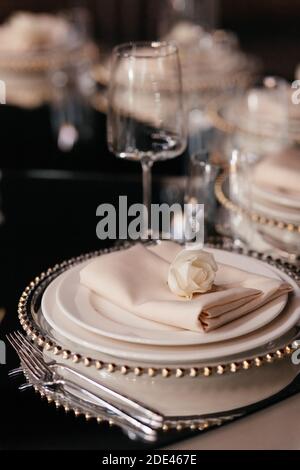 White rose on the set of plates on the dark background. Luxury table set in the restaurant. Glasses and plates. Wedding banquet.  Stock Photo
