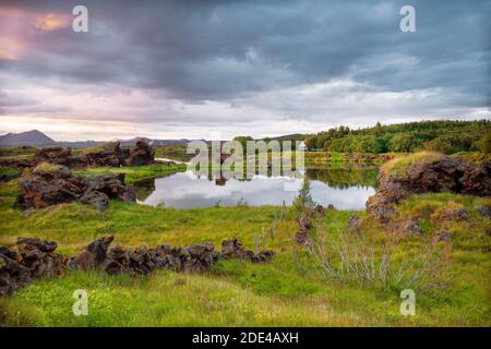 Lava formations of volcanic rock rise from green meadows with dramatic skies at Kalfastroend, house is reflected in lake Myvatn, Skutustaoir Stock Photo