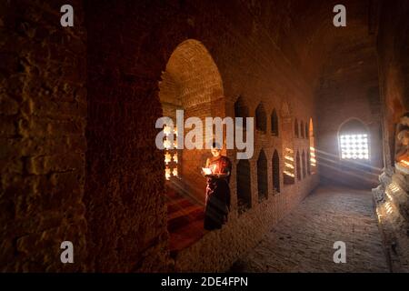 Buddhist young monk in red robe reading standing in front of rays of light in a temple, Bagan, Myanmar Stock Photo