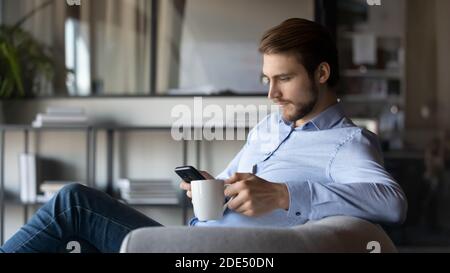 Pensive young businessman focused on smartphone holding cup of tea Stock Photo