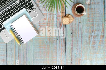 Computer laptop, face mask, coffee and supplies on wood table. Top view. Work from home during pandemic concept. Stock Photo