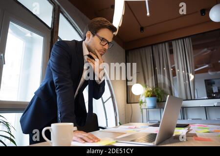 Male executive standing by table studying data on laptop screen Stock Photo
