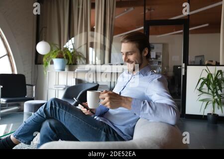 Smiling man employee enjoying hot drink in office using cellphone Stock Photo