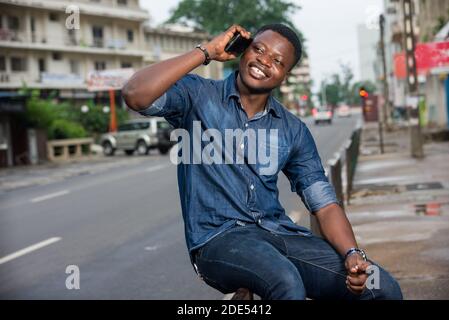 Portrait of happy young man sitting outside looking to the side while talking on smart phone outdoors in the street. Stock Photo