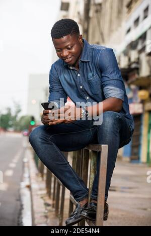 Portrait of a smiling young man sitting with a mobile phone on an iron bar in the street Stock Photo