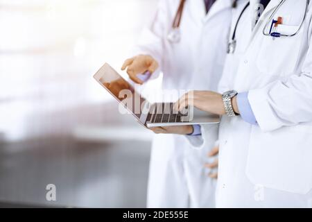 Two unknown doctors with stethoscopes discuss medical exam resoults, while standing at sunny hospital office. Physicians using a laptop for checking Stock Photo