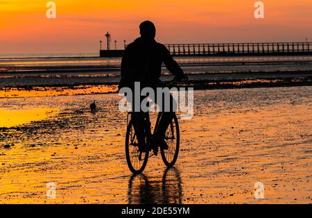 A male cyclist takes a ride on a bicycle on the beach on a Winter's evening as the sun sets at low tide in Littlehampton, West Sussex, England, UK. Stock Photo