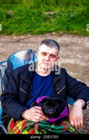 Man tourist sitting smiling with dog black pug in touristic chair under checkered plaid in nature Stock Photo
