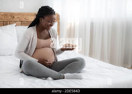 happy pregnant woman with plate of cookies sitting in bed at home Stock Photo