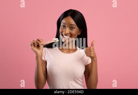 Pretty black lady with beautiful smile holding wooden toothbrush and showing thumb up gesture on pink background Stock Photo