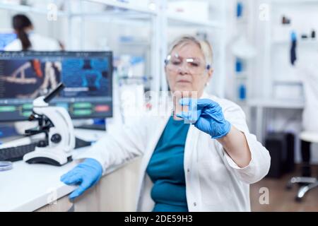 Senior scientist in protective gear holding sample during clinic study giving expertise. Elderly researcher in sterile lab looking on microscope slide wearing lab coat. Stock Photo