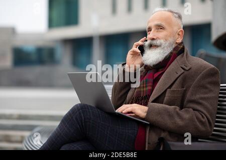 Elderly businessman with laptop sitting on bench, talking on phone Stock Photo