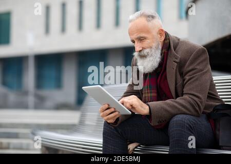 Stylish grey-haired man sitting on the bench, using digital tablet Stock Photo