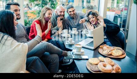 Beautiful young friends waving hand and talking while having a video call with their friends on a laptop in a coffee shop with face mask down - Studen Stock Photo